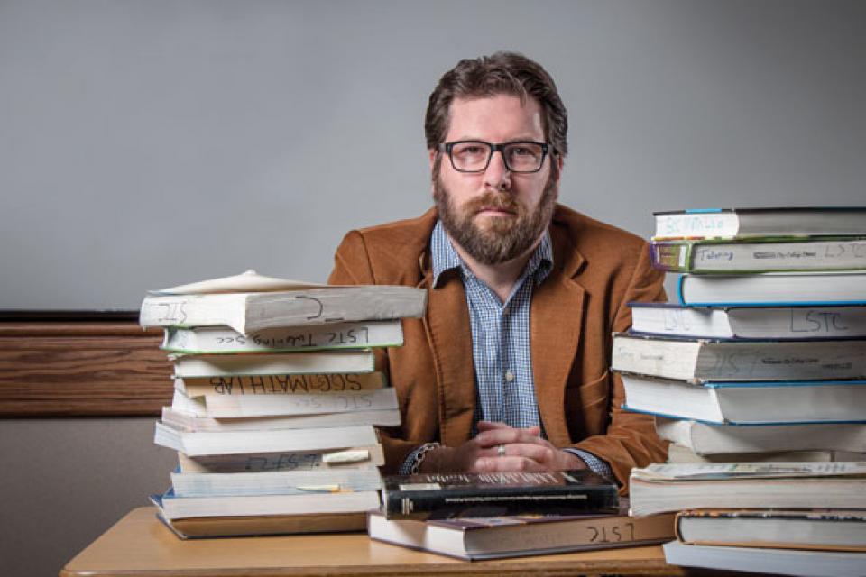 Paul Baltimore photo with stacks of books in front of him at teacher's desk